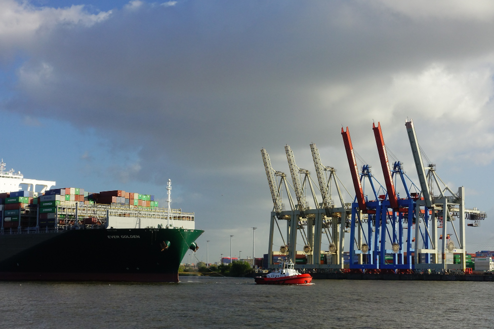 Large container ship “EVER GOLDEN” approaching a port with colorful cargo cranes and a tugboat assisting in docking operations.