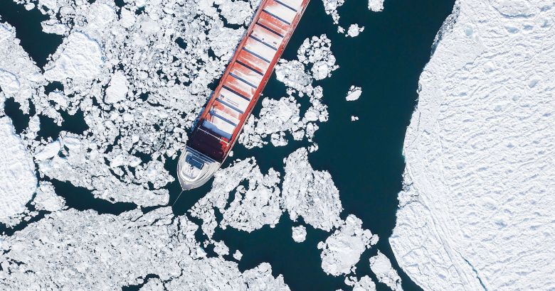 Aerial view of a large cargo ship navigating through icy waters surrounded by floating ice chunks and a snowy coastline.